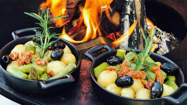 Kleine Pfännchen mit Kartoffeln, Gemüse, Oliven und getrockneten Tomaten auf der Grillfläche des swissfirecube. Dahinter das brennende Feuer